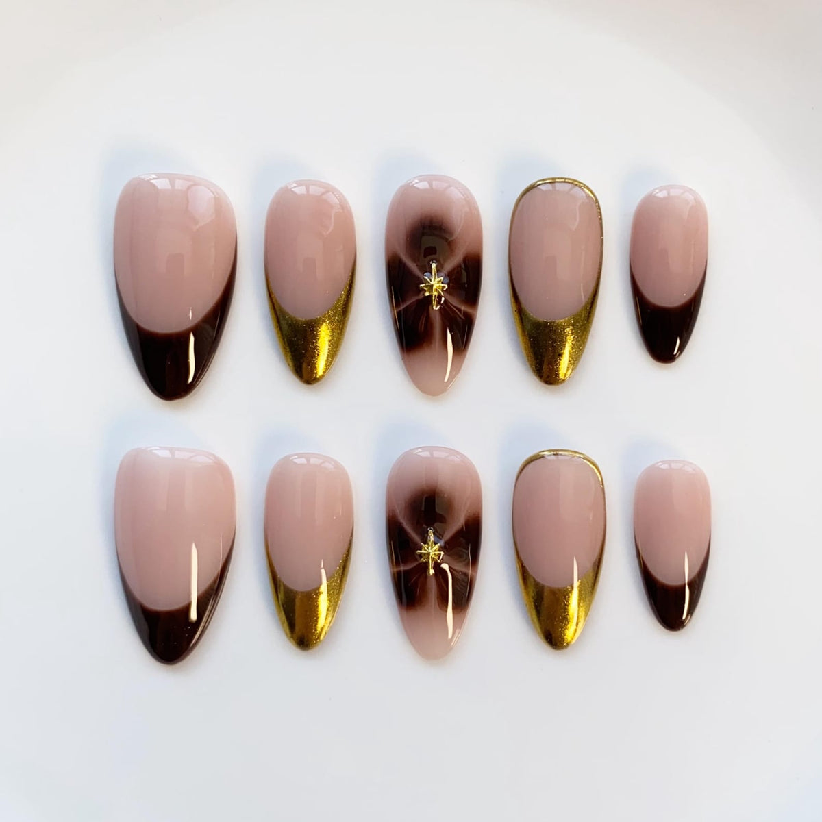 A set of ten artificial nails in various sizes. The nails feature a nude base with dark brown tips, gold accents, and a brown and gold starburst design. Some nails have a gold border.