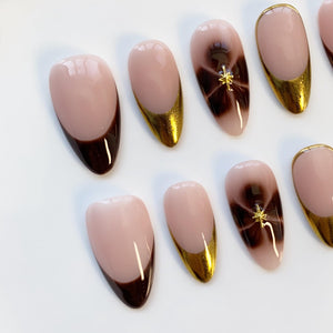 Oval-shaped artificial nails in various designs. Some nails feature a dark brown French tip, while others have a gold glitter tip. A few nails have a brown swirl design with a gold star accent. The nails are set against a white background.
