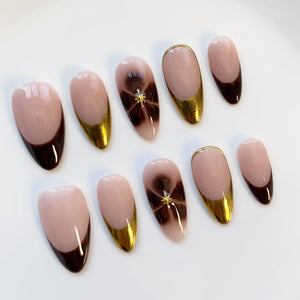 A set of ten artificial nails in various designs. Some nails feature a nude base with a dark brown tip, while others have a gold metallic edge. Two nails have a brown swirl design with a gold star accent.