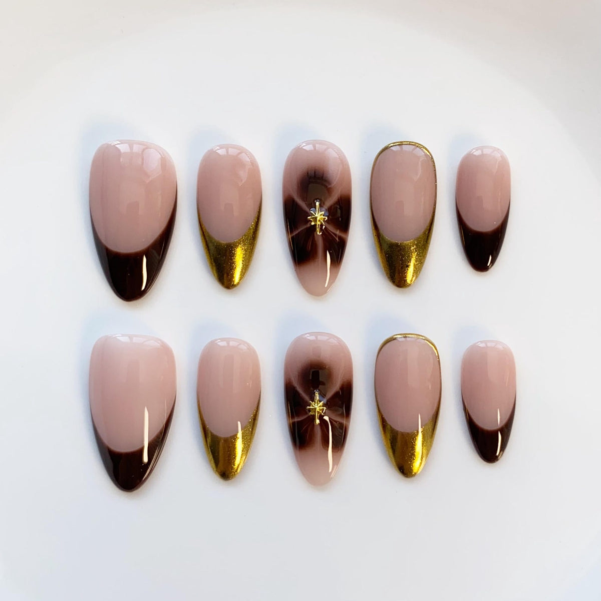 A set of ten artificial nails in various shapes. The nails feature a light pink base with dark brown tips, gold accents, and a dark brown swirl design with a gold star. The nails are arranged on a white surface.