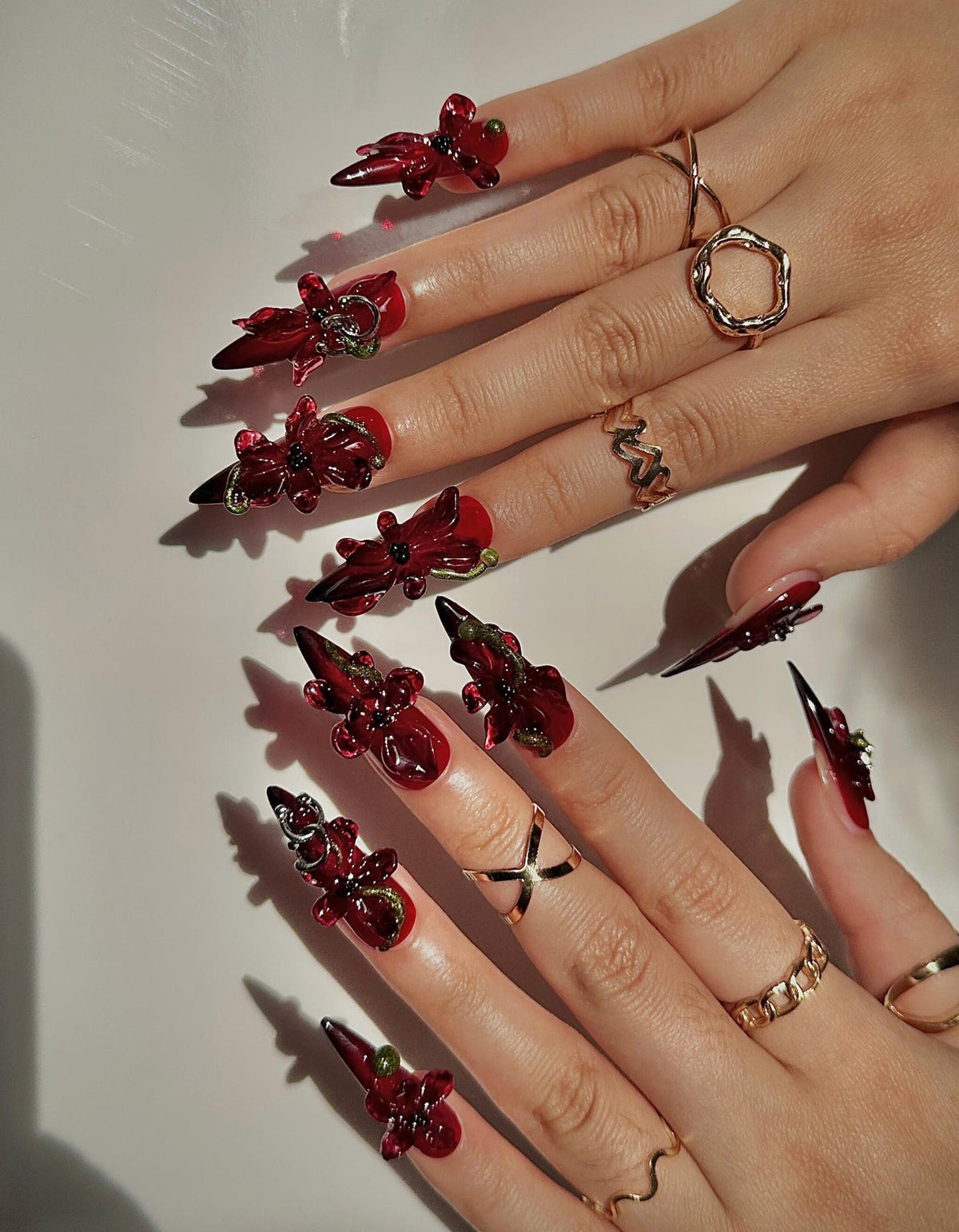A close-up of hands showcasing elaborate, long, red and black nail art. Each nail features a glossy red base with intricate floral-like embellishments and pointed black tips. Gold rings of various designs adorn the fingers, complementing the nail art