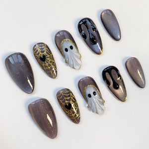A set of Halloween-themed nail art designs. The nails feature various designs, including a gold glitter spiderweb with a black heart, a white ghost, and black dripping paint. The nails are in shades of gray and gold.