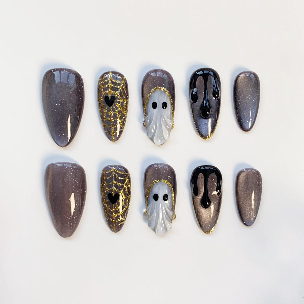 A set of ten press-on nails in various designs. Some nails are a shimmery gray, others feature gold spiderwebs with black hearts, white ghost designs, and black dripping paint. The nails are oval-shaped.