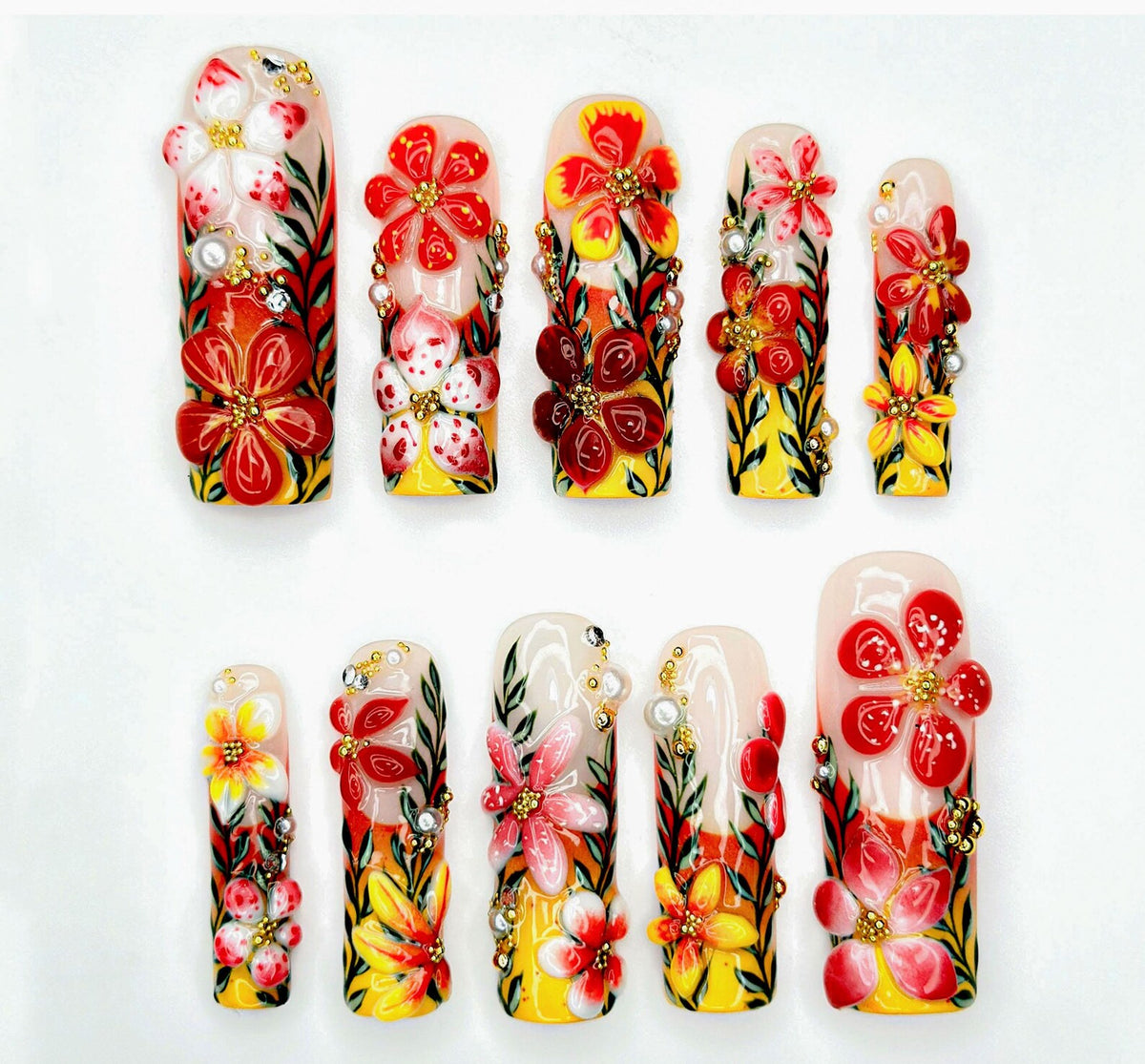 A set of ten artificial nails with a floral design. Each nail features a base of yellow, orange, and red, with 3D flowers in red, white, and pink. Some nails have pearl and rhinestone accents.