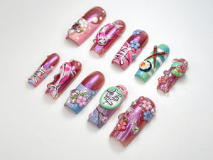 A collection of elaborately designed artificial nails. Each nail features unique artwork, including floral patterns, koi fish, and Japanese text. Colors include pink, purple, blue, and green, with embellishments like rhinestones and pearls.