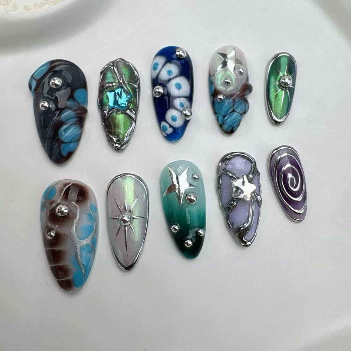 A collection of ten uniquely designed artificial nails. Each nail features a different design, including swirls, stars, and abstract patterns, with a variety of colors such as blue, green, and purple. Some nails have silver embellishments.