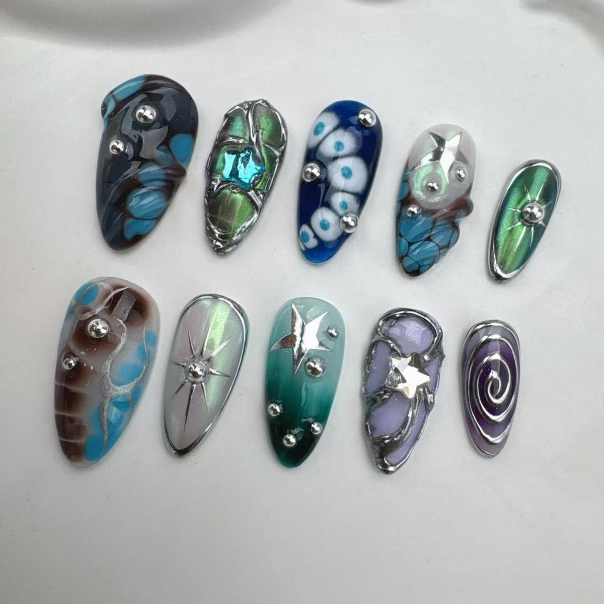 A collection of ten uniquely designed artificial nails. Each nail features a different design, including swirls, floral patterns, and geometric shapes, with accents of silver and various colors like blue, green, and purple. The nails are arranged on 