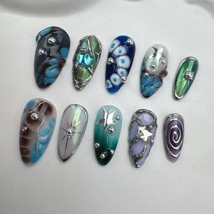 A collection of ten uniquely designed artificial nails. Each nail features a different design, including swirls, floral patterns, and geometric shapes, with accents of silver and various colors like blue, green, and purple. The nails are arranged on 