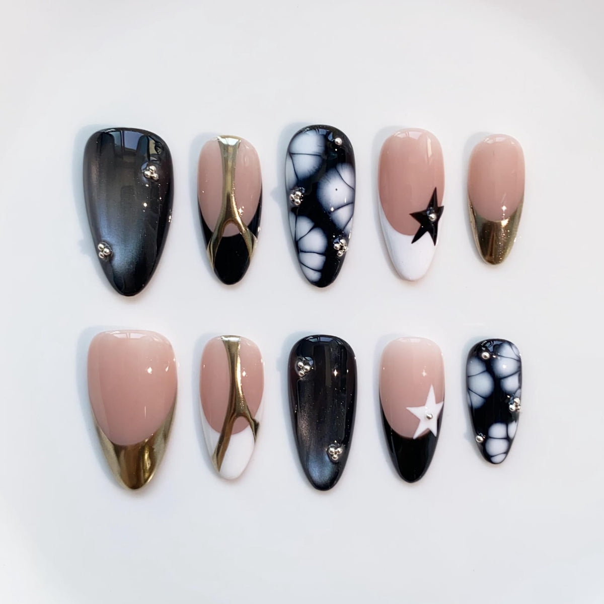 A set of ten artificial nails in various designs. The nails feature a mix of black, gold, white, and nude colors. Some nails have star and floral patterns, while others have metallic accents.