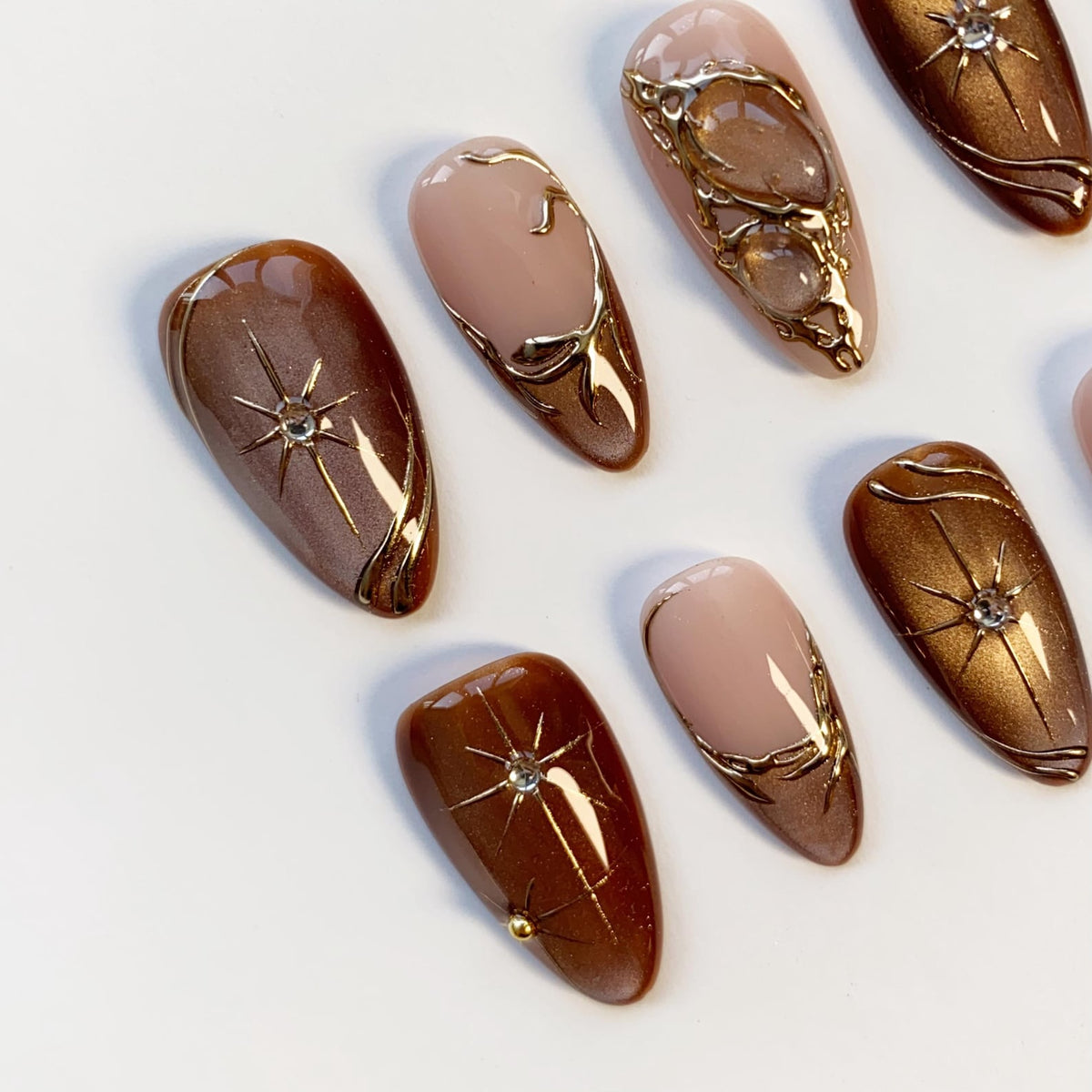 A collection of almond-shaped false nails in various shades of brown and nude. Some nails feature gold metallic accents, including starburst designs and ornate swirls. Several nails are adorned with small, sparkling rhinestones.