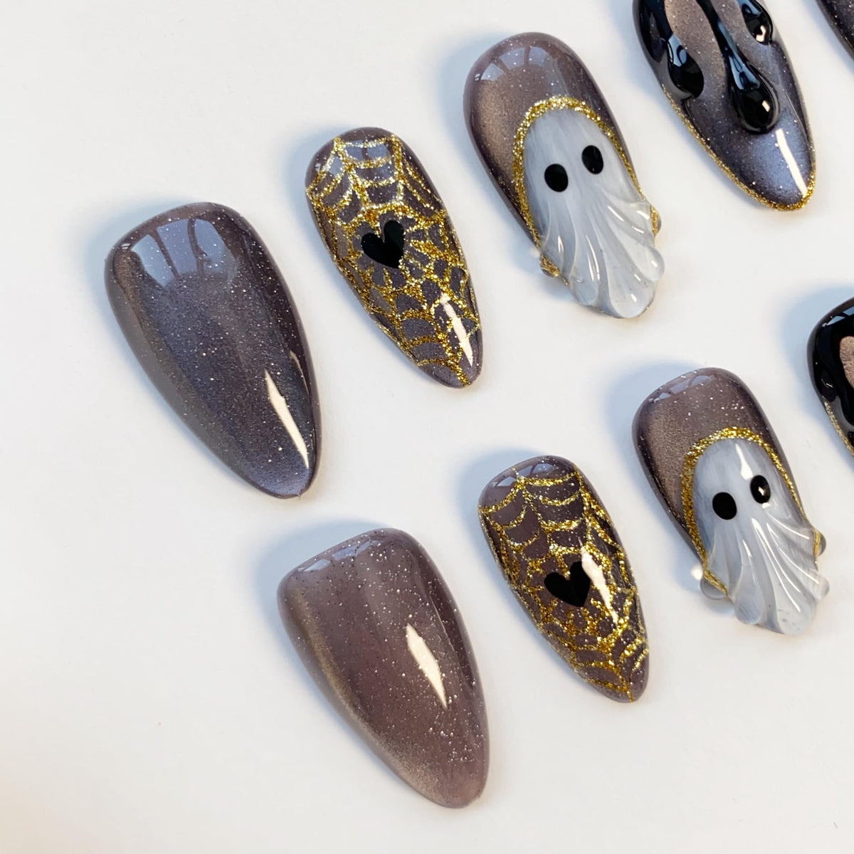 A collection of Halloween-themed false nails. Designs include glittery purple, gold spiderwebs with black hearts, and ghostly white figures with black eyes. The nails are almond-shaped.