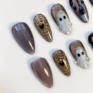 A collection of Halloween-themed false nails. Designs include glittery purple, gold spiderwebs with black hearts, and ghostly white figures with black eyes. The nails are almond-shaped.