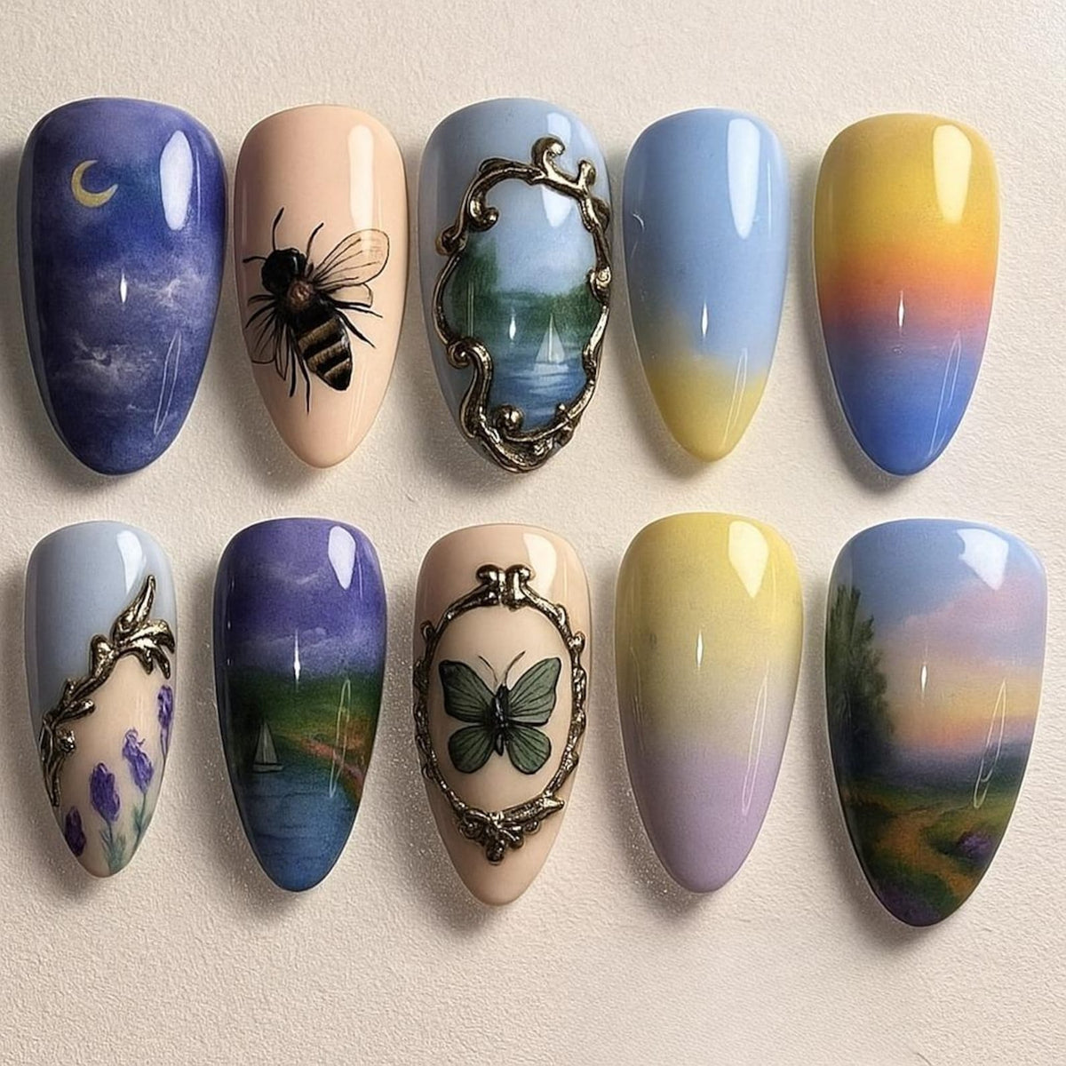 A set of ten painted artificial nails. Designs include a night sky with a crescent moon, a bee, a landscape scene, and a butterfly. Colors include blue, yellow, beige, and gold.