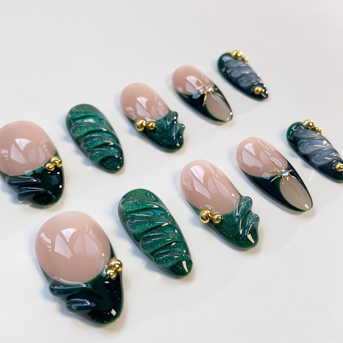 A set of ten artificial nails in various designs. Some nails feature a nude base with a dark green and gold accent, while others have a dark green textured design. Several nails have gold embellishments.