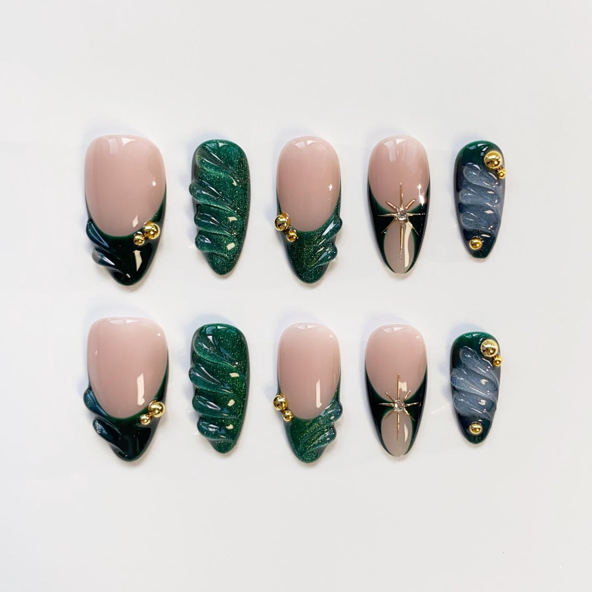 A set of ten artificial nails in various designs. The nails feature a nude base with dark green and gold accents, along with a few nails with a silver star design and a swirling blue-gray pattern. The nails are oval-shaped.