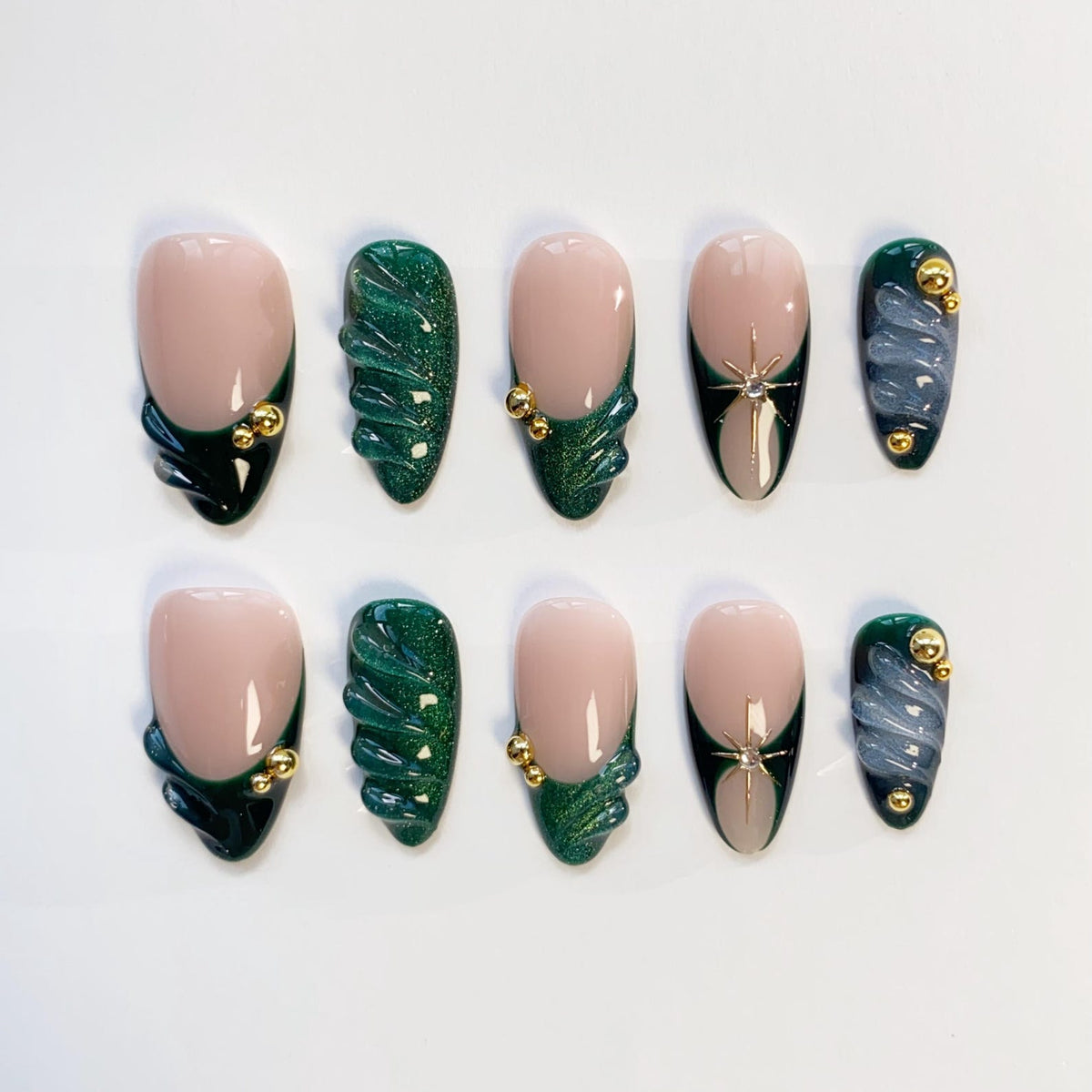 A set of ten artificial nails in various designs. The nails feature a nude base with dark green and gold accents, including a textured wave design and starburst embellishments. Some nails have a dark green French tip.