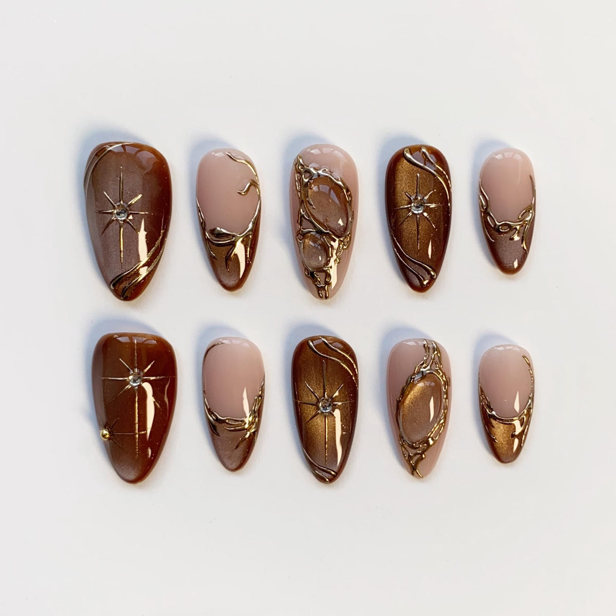A set of ten almond-shaped press-on nails. The nails feature a combination of brown, nude, and gold colors. Some nails have starburst and swirl designs, while others have gold embellishments. The nails are arranged on a white surface.