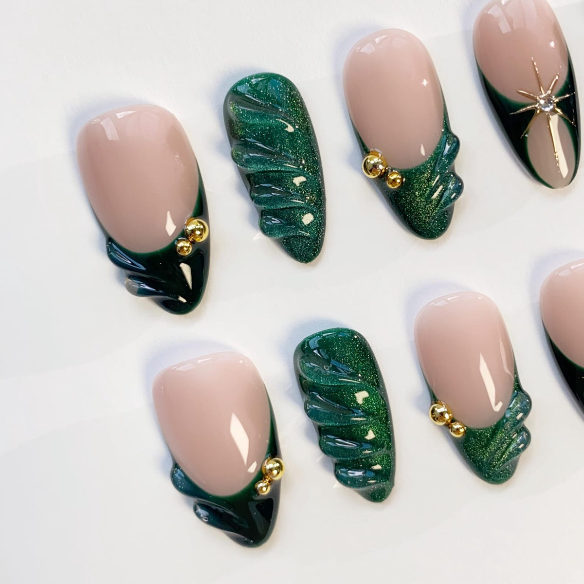 Oval-shaped artificial nails with a nude base and dark green accents. Some nails feature a textured, glittery green design, while others have gold embellishments. A star-shaped jewel adorns one nail.