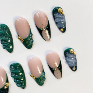 A collection of artificial nails in various designs. Some nails feature a nude base with a dark green and gold accent, while others have a dark green wing-like design. Several nails have gold embellishments and a star-shaped jewel.