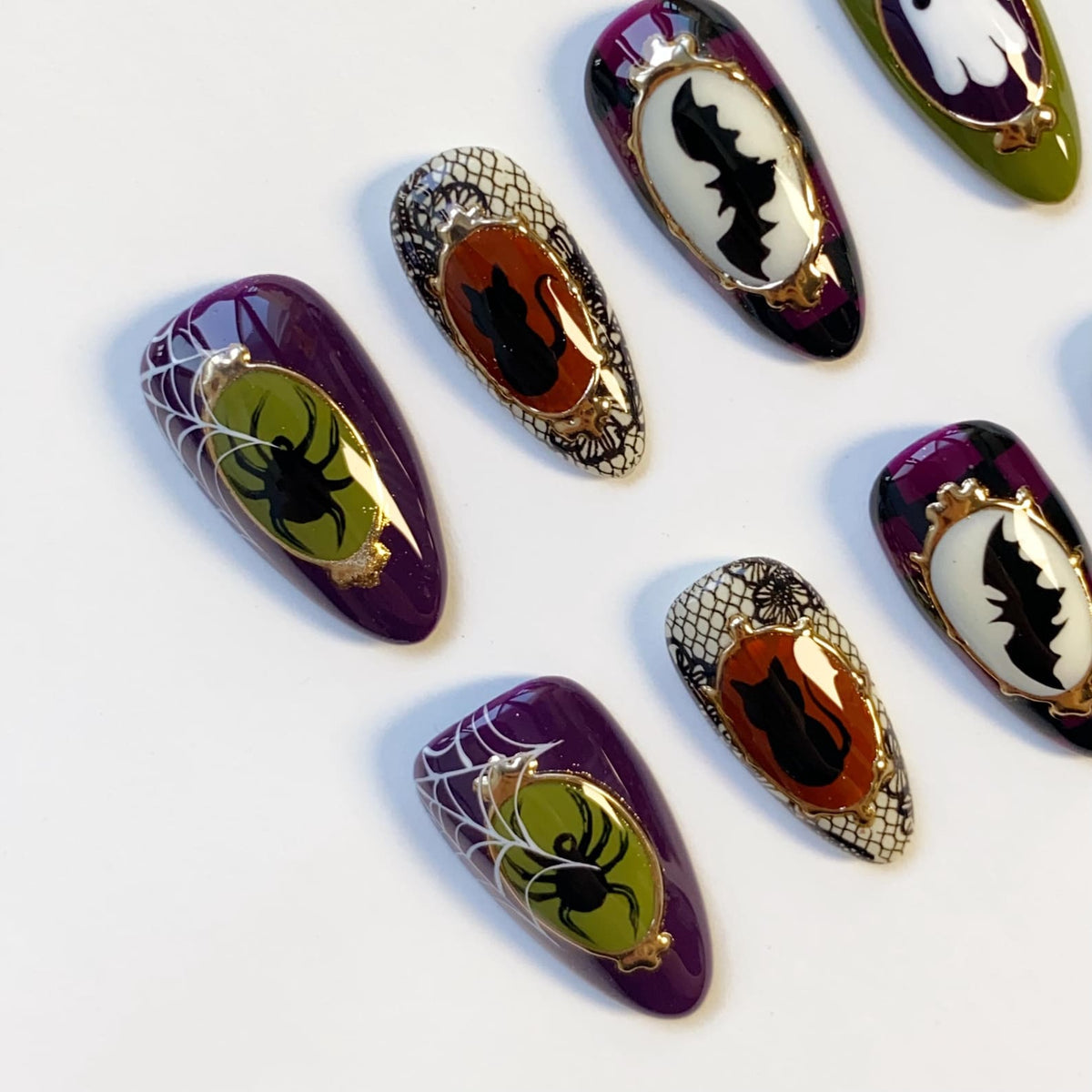 A collection of Halloween-themed false nails. The nails feature designs of spiders, bats, and black cats in ornate frames, with a color palette of purple, black, white, and gold. Some nails have a spiderweb design.