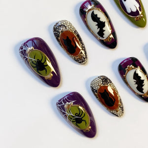 A collection of Halloween-themed false nails. The nails feature designs of spiders, bats, and black cats in ornate frames, with a color palette of purple, black, white, and gold. Some nails have a spiderweb design.