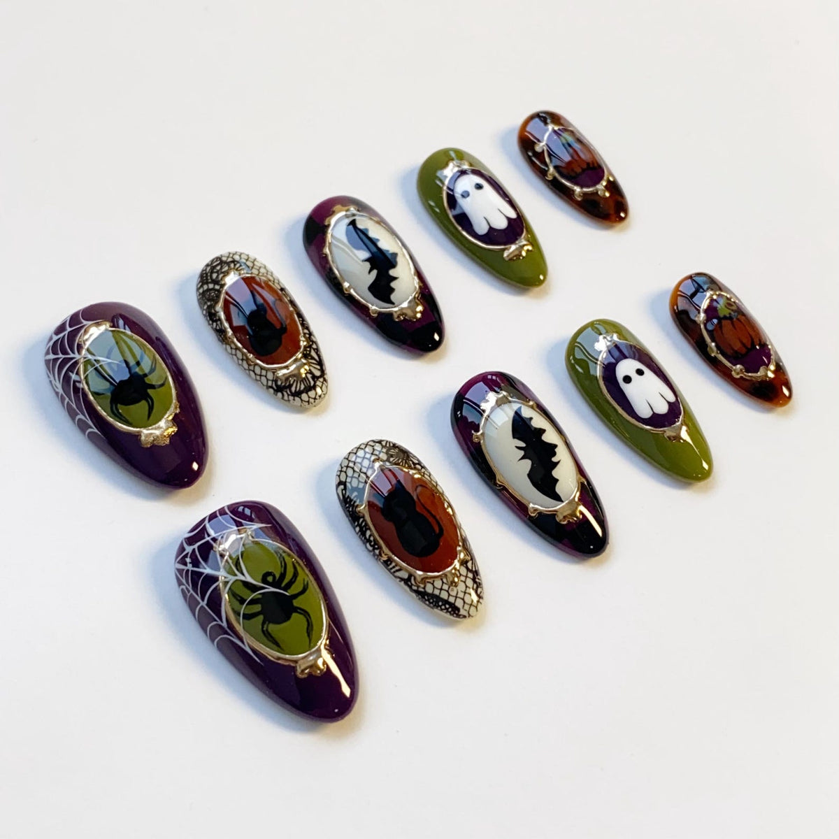 A collection of Halloween-themed press-on nails. The nails feature designs such as spiders, bats, ghosts, and pumpkins. Colors include purple, green, orange, and black, with gold accents. The nails are oval-shaped and have a glossy finish.