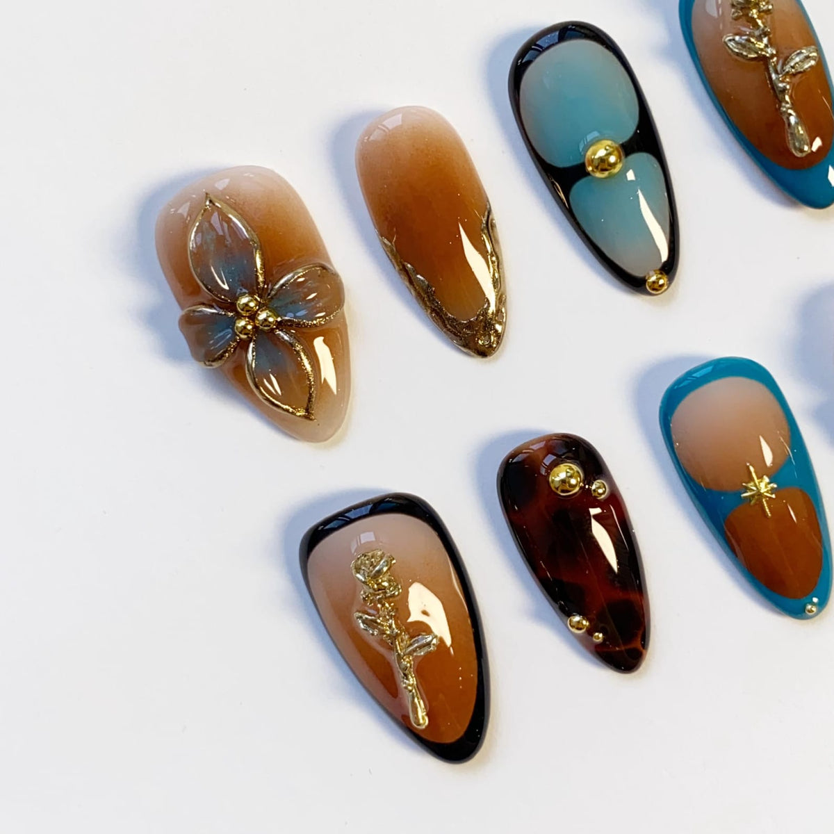 A collection of artificial nails with various designs. Some nails feature gold floral embellishments, while others have abstract patterns in shades of brown, blue, and black. The nails are oval-shaped and have a glossy finish.