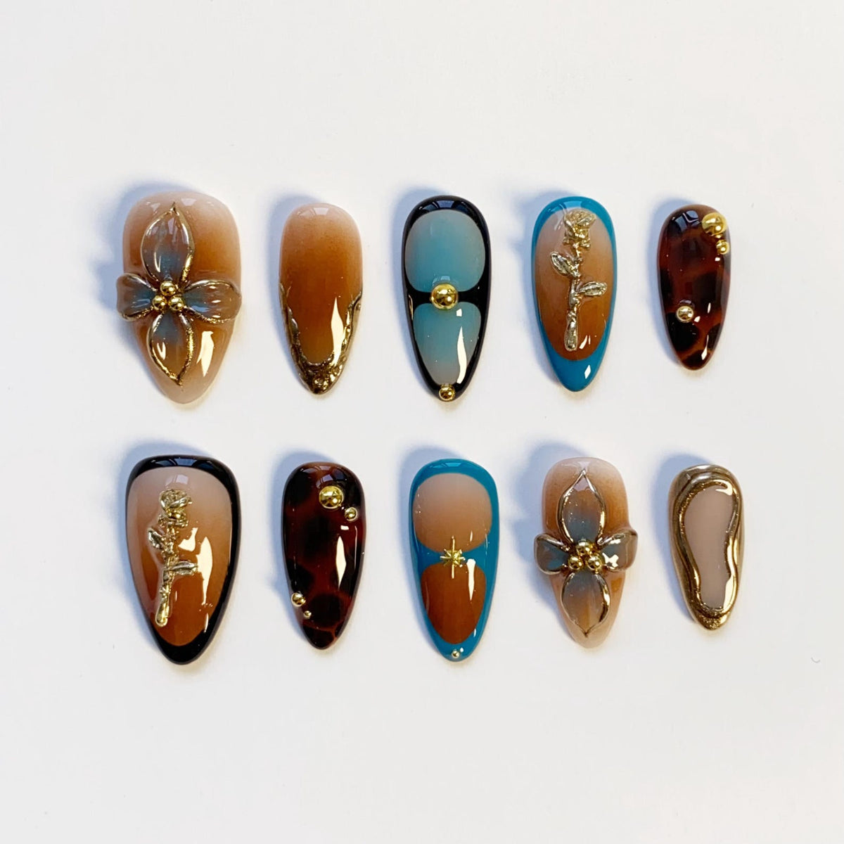 A set of ten artificial nails in various designs. The nails feature a range of colors, including brown, beige, and blue, with gold accents. Some nails have floral or abstract designs, while others have a tortoiseshell pattern.
