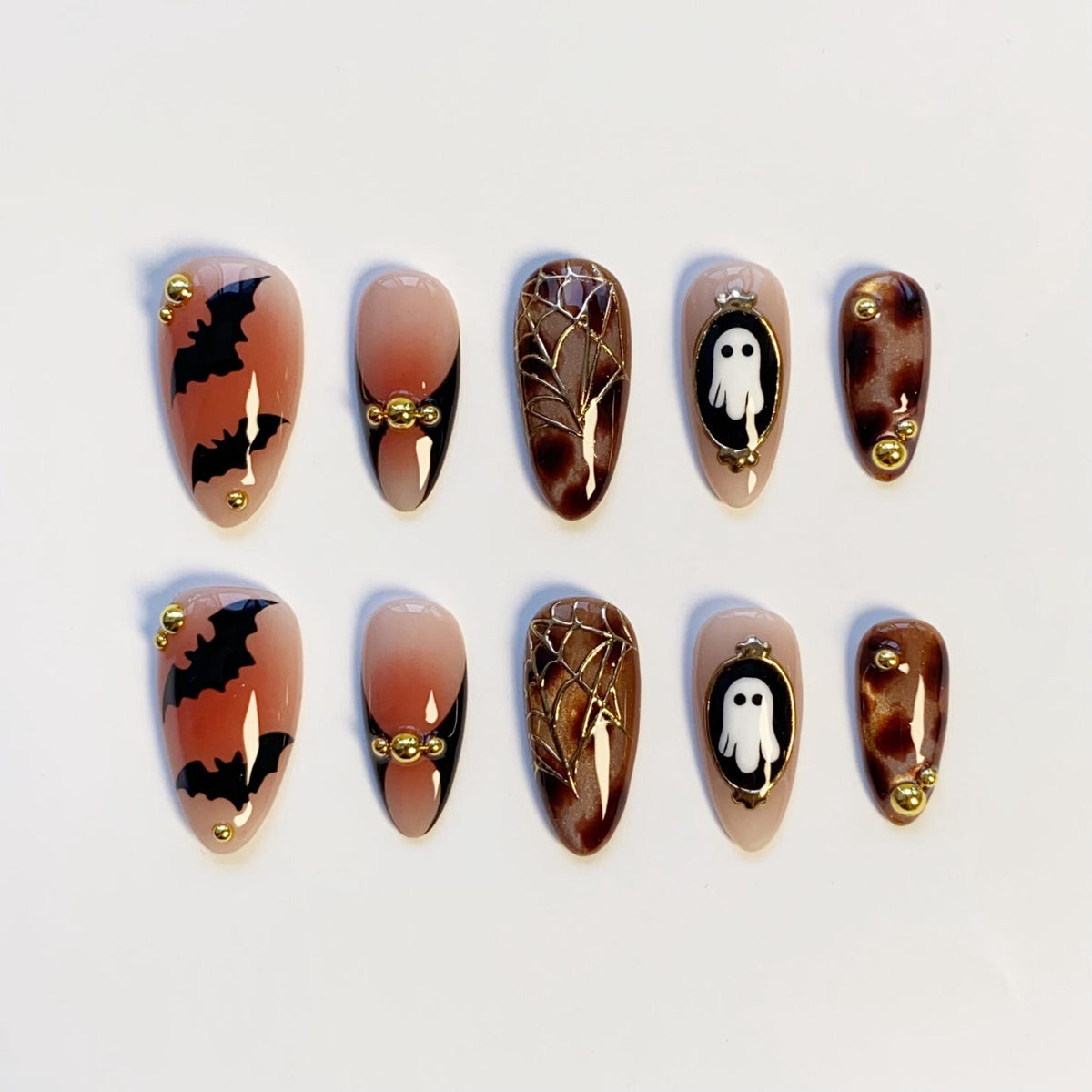 A set of ten almond-shaped press-on nails with a Halloween theme. Designs include bats, spiderwebs, ghosts, and a gradient effect. The nails are in shades of nude, brown, and black, with gold accents.