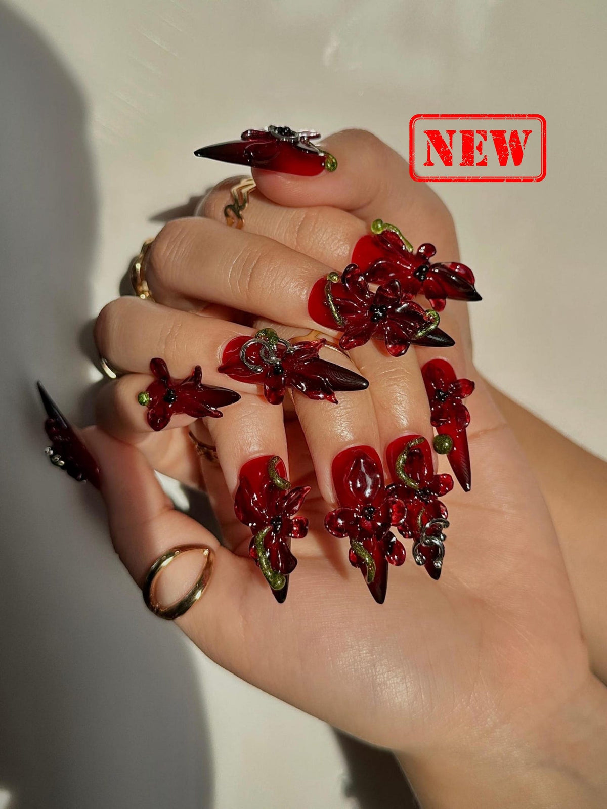 A hand displays long, red and black stiletto nails adorned with 3D floral designs. The nails have a gradient effect, transitioning from red to black at the tips. The word "NEW" is stamped in a red rectangle.