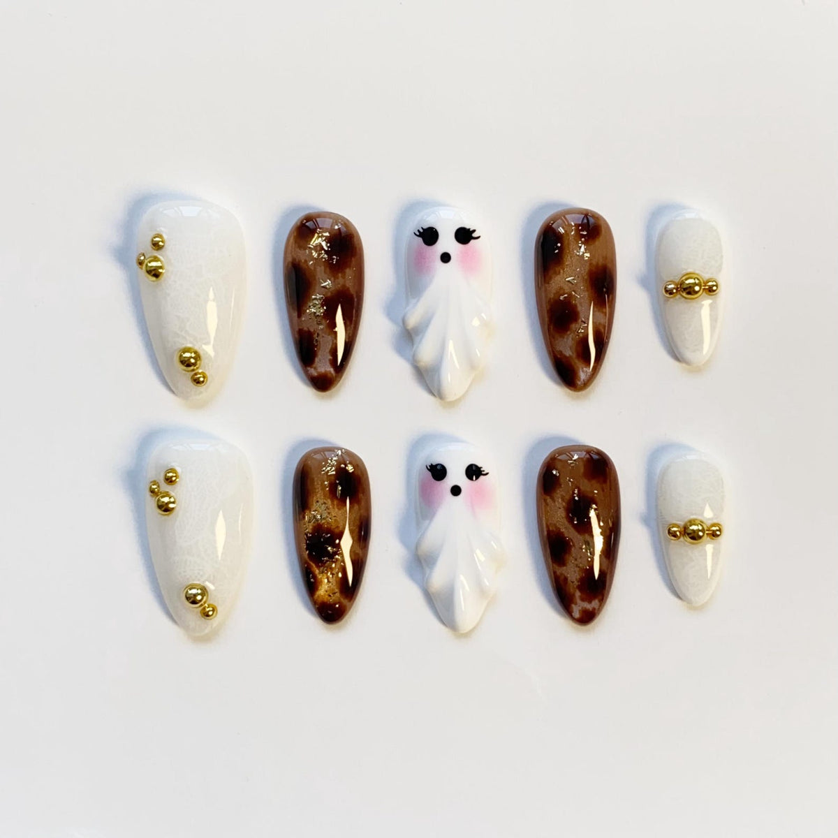 A set of ten press-on nails. The nails feature a variety of designs, including white with gold accents, brown leopard print, and white ghost designs with pink cheeks. The nails are oval-shaped.