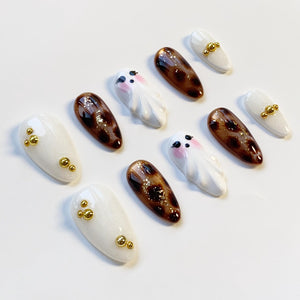 A collection of artificial nails in various designs. Some nails are white with gold embellishments, others feature a brown leopard print, and some are shaped like cartoon ghosts with pink cheeks and black eyes.