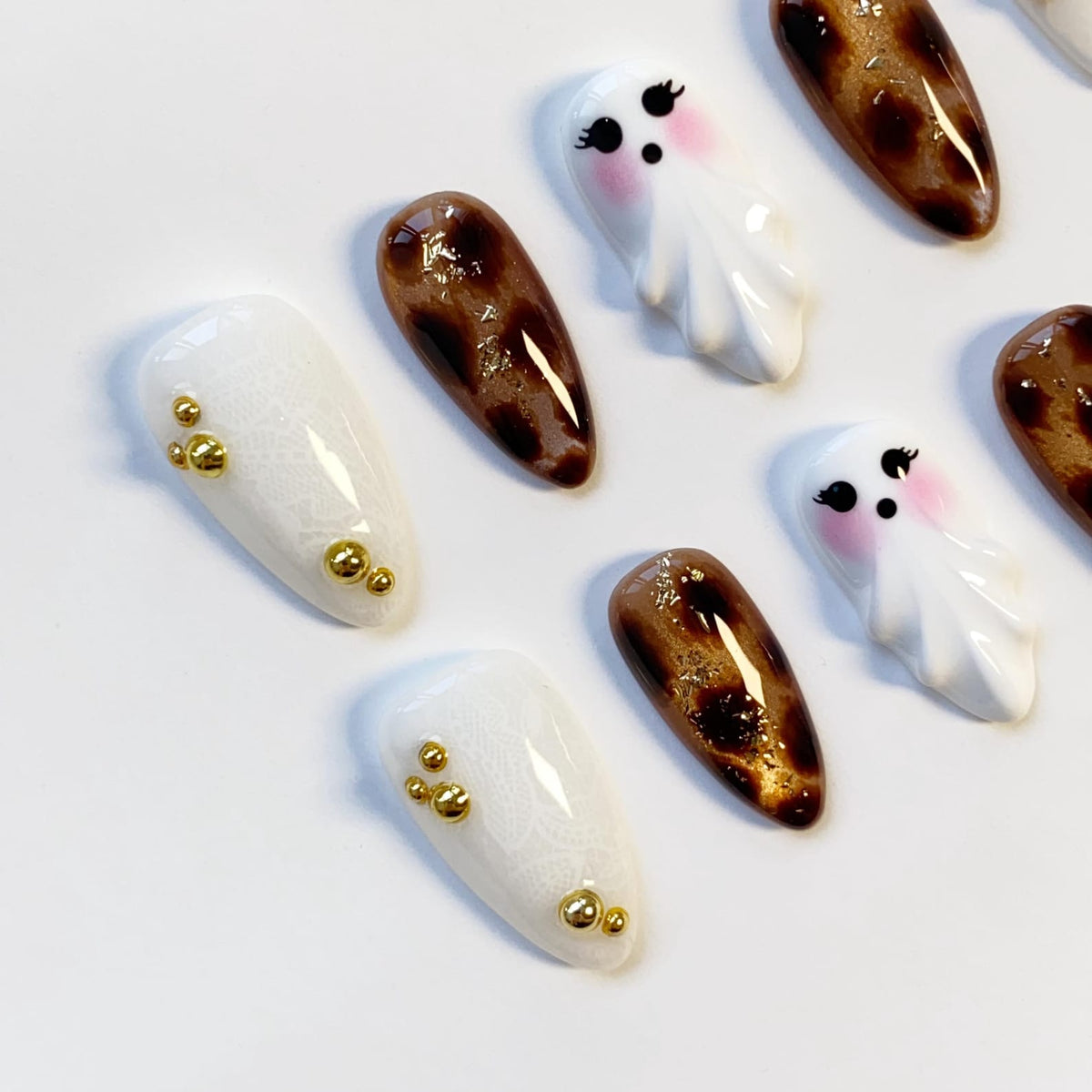 A collection of artificial nails with various designs. Some nails are white with gold embellishments, others feature a brown and gold pattern, and some are shaped like cartoon ghosts with pink cheeks.