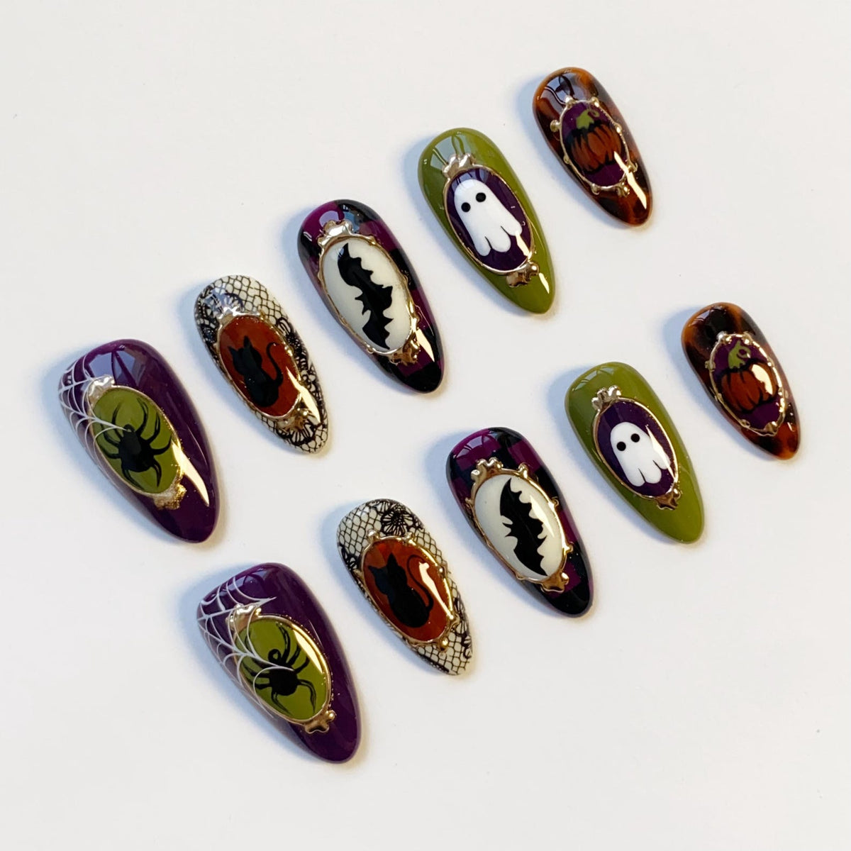 A collection of Halloween-themed false nails. Designs include a spider web with a spider, a bat, a ghost, and a pumpkin. The nails feature a variety of colors, including purple, green, orange, and black, with gold accents.