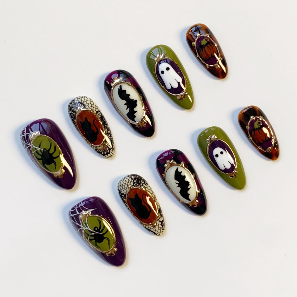 A collection of Halloween-themed false nails. Designs include a spider web with a spider, a bat, a ghost, and a pumpkin. The nails feature a variety of colors, including purple, green, orange, and black, with gold accents.