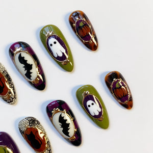 Halloween-themed nail art featuring various designs. Some nails have a white ghost, a black bat, or a pumpkin in a gold frame. The nails are painted in shades of green, purple, and orange.