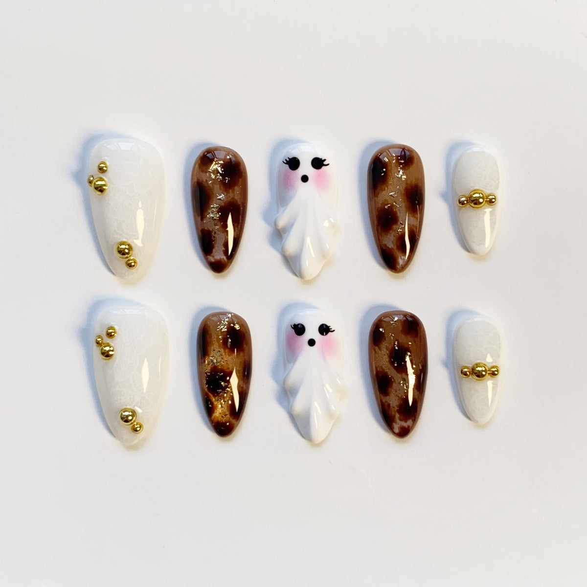A set of ten press-on nails in various designs. The nails feature white, brown leopard print, and ghost designs. Some nails have gold bead accents. The nails are oval-shaped and are arranged on a white surface.