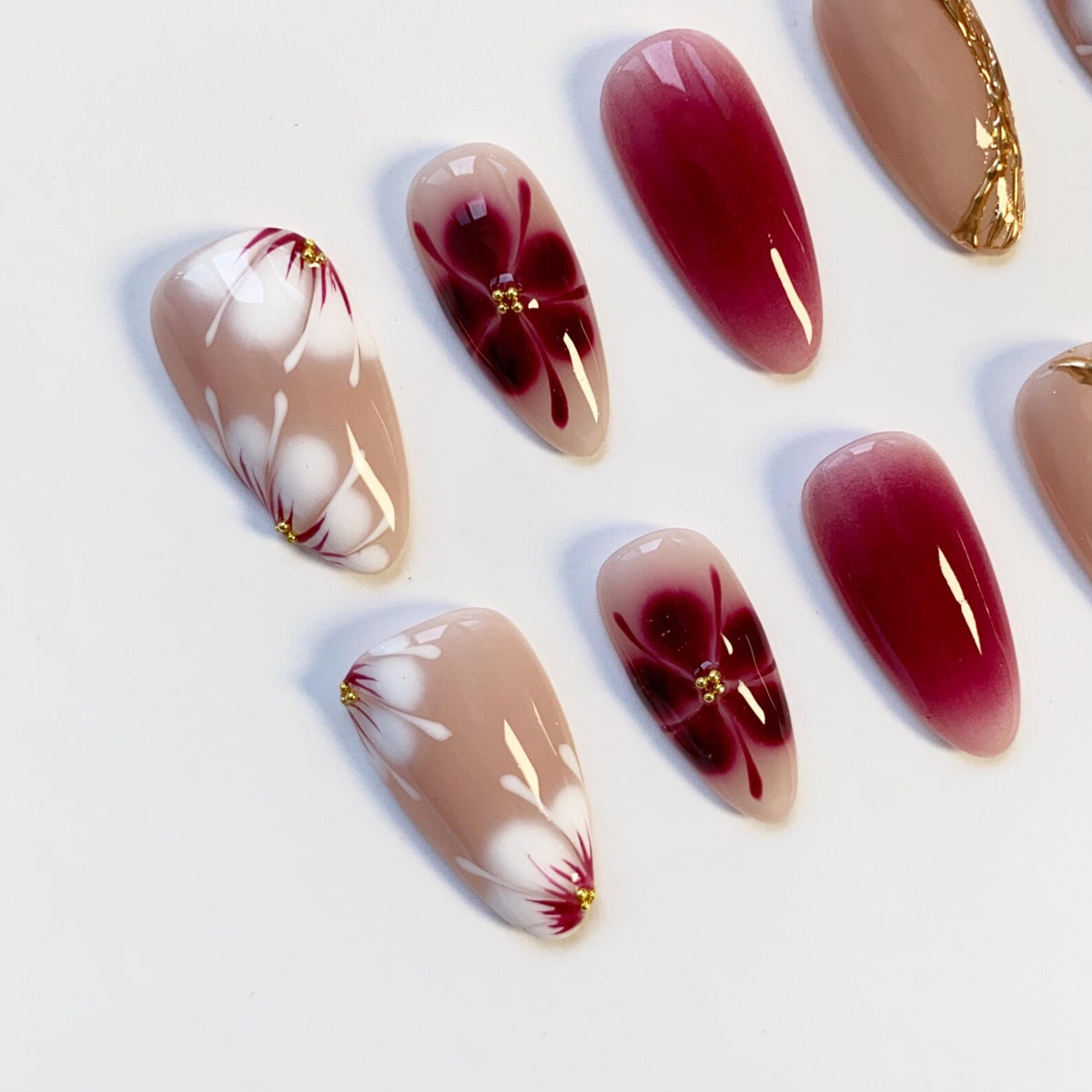 A collection of almond-shaped artificial nails. Some nails feature a gradient of red to pink, while others showcase a floral design with white petals, red accents, and gold embellishments. The nails are set against a white background.