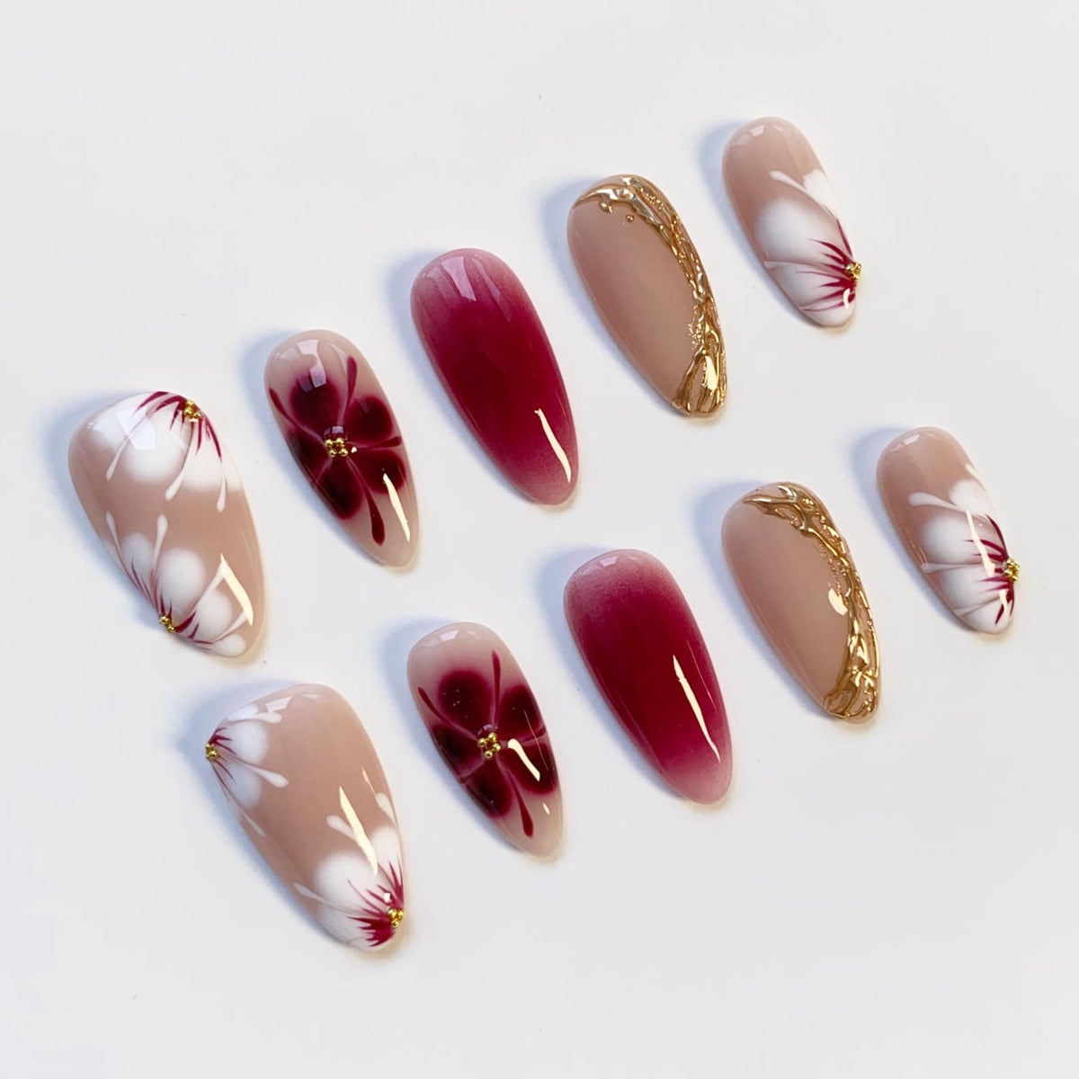 A set of ten artificial nails with a variety of designs. Some nails are nude with white floral patterns and gold accents. Others feature a deep red ombre effect or gold filigree. The nails are oval-shaped.