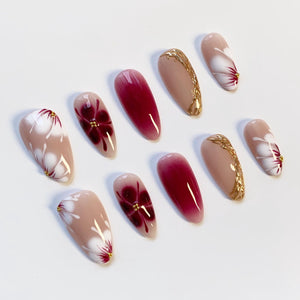 A set of ten artificial nails with a variety of designs. Some nails are nude with white floral patterns and gold accents. Others feature a deep red ombre effect or gold filigree. The nails are oval-shaped.
