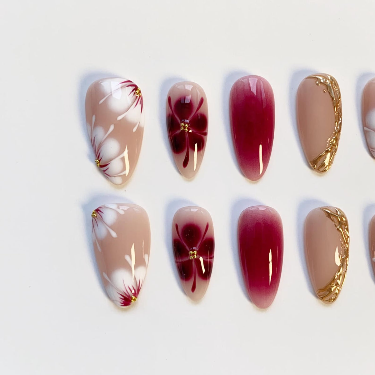 A set of almond-shaped false nails. Some nails feature a nude base with white floral designs and gold accents. Others have a deep red gradient or a burgundy flower design with gold details. The nails are arranged on a white surface.