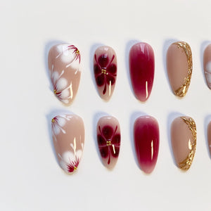 A set of almond-shaped false nails. Some nails feature a nude base with white floral designs and gold accents. Others have a deep red gradient or a burgundy flower design with gold details. The nails are arranged on a white surface.