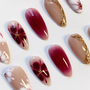 A collection of artificial nails in various designs. Some nails feature a gradient from light to dark red, while others showcase floral patterns in burgundy and gold accents. The nails are oval-shaped and have a glossy finish.