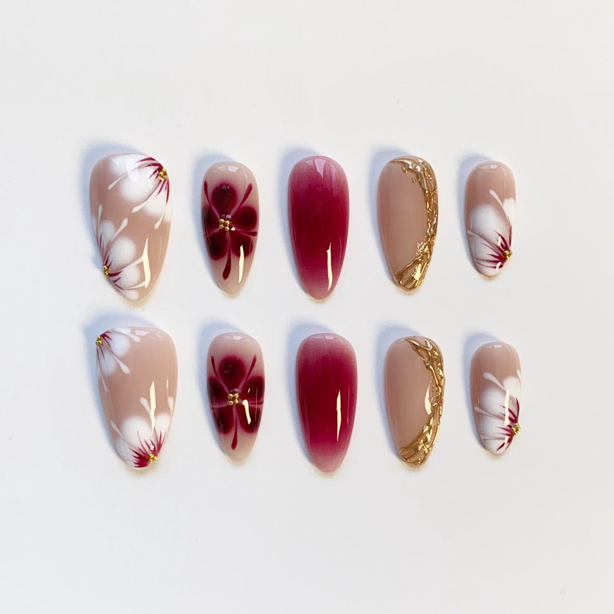 A set of ten artificial nails in various designs. Some nails are nude with white floral designs and gold accents. Others are a deep red gradient or nude with gold decorative lines. The nails are oval-shaped.