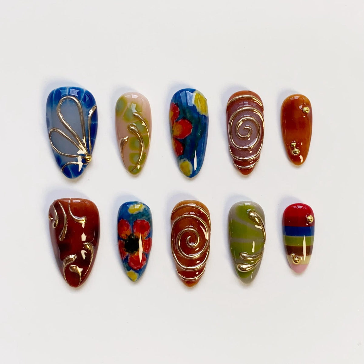 A collection of ten almond-shaped, painted artificial nails. Each nail features unique designs, including floral patterns, swirls, and gold accents. Colors range from blue and red to brown and green, with a glossy finish.