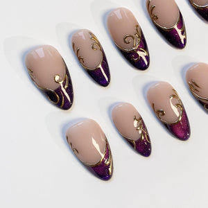 Oval-shaped artificial nails with a nude base and a deep purple glittered lower section. Gold filigree designs adorn the upper portion of each nail, creating an elegant and ornate appearance. The nails are arranged on a white surface.