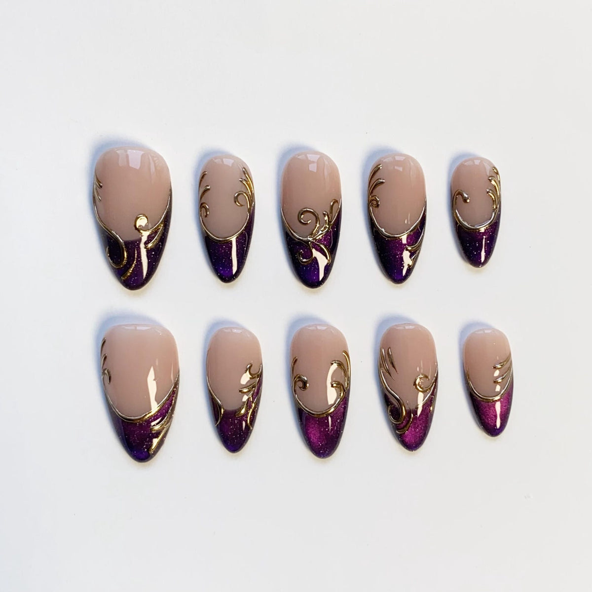 Set of ten almond-shaped false nails. Each nail features a nude base, a deep purple glitter accent at the base, and gold filigree detailing. The nails are arranged on a white surface.