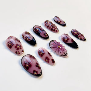 A set of ten artificial nails in various designs. The nails are primarily shades of pink and purple, with gold accents. Some nails feature a floral pattern, while others have a black tip.