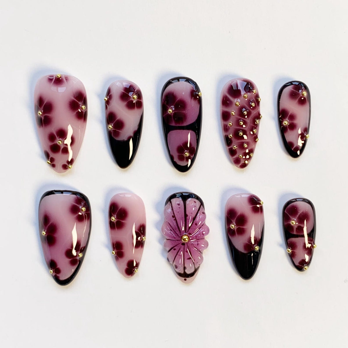A set of ten artificial nails with a floral design. The nails are primarily pink with dark red flower accents and small gold studs. Some nails have a black tip.