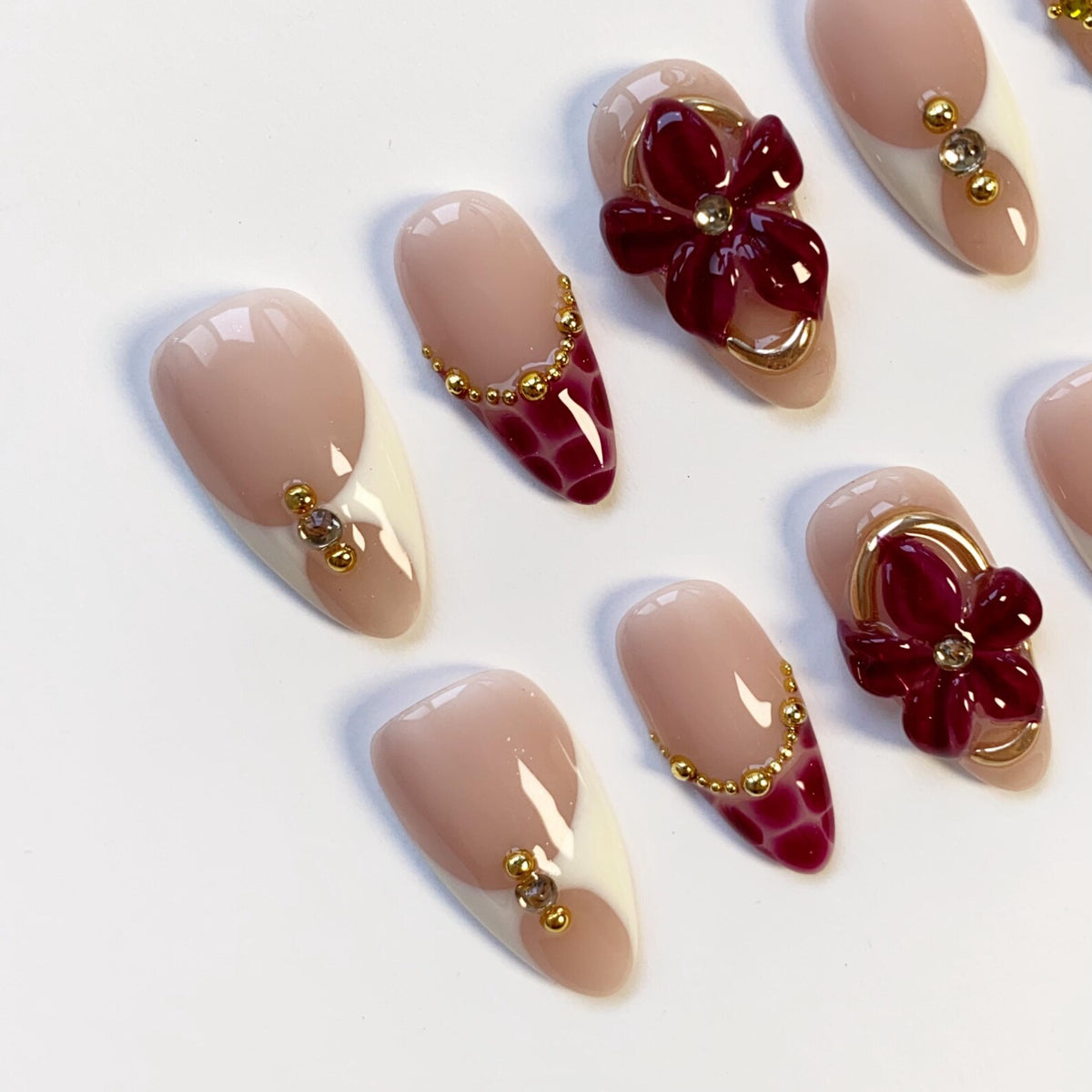 A collection of almond-shaped artificial nails. Some nails feature a two-tone design with a nude base and white tips. Others have a burgundy pattern or a 3D flower design, all adorned with gold-colored embellishments.